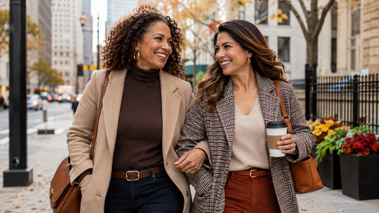 Two women walking arm in arm on a city sidewalk during autumn. The woman on the left wears a beige blazer over a dark brown turtleneck, and the woman on the right wears a brown and beige plaid blazer over a light beige top.